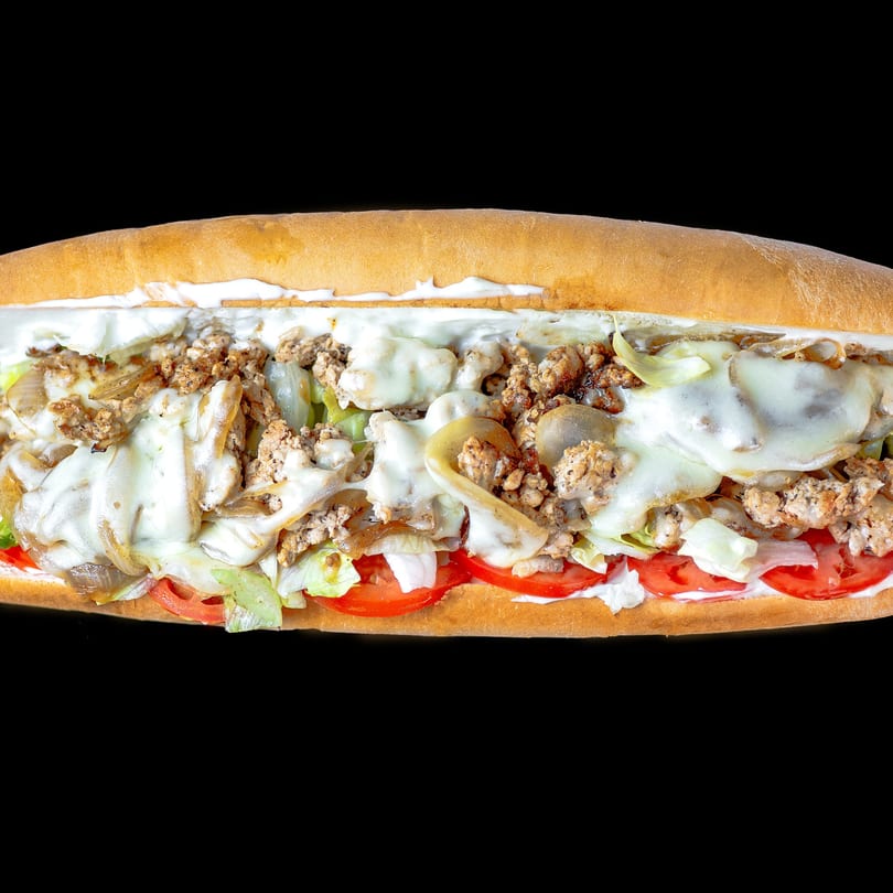 A chicken cheesesteak sub with melted cheese, lettuce, tomatoes, and onions on a long roll.
